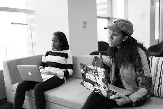 Two women seated indoors with laptops, engaged in discussion.